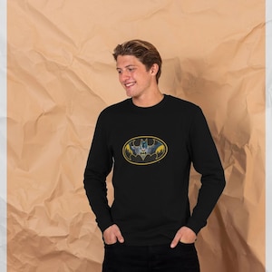 May include: Black long-sleeve shirt featuring the Batman logo in a yellow oval. The logo depicts a bat silhouette with a cityscape background. The shirt is made of a soft material and is suitable for casual wear.