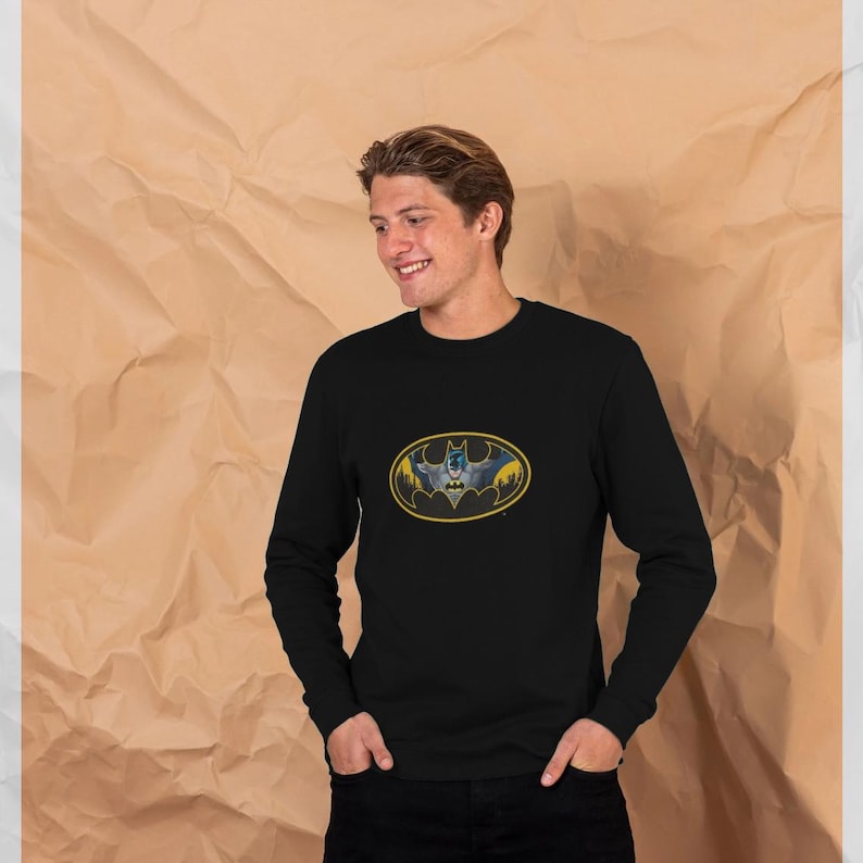 May include: Black long-sleeve shirt featuring the Batman logo in a yellow oval. The logo depicts a bat silhouette with a cityscape background. The shirt is made of a soft material and is suitable for casual wear.
