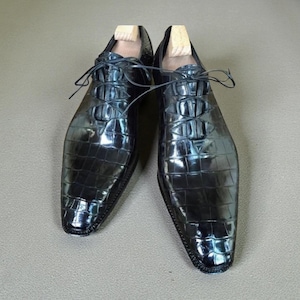 May include: A pair of black, glossy leather dress shoes with a textured, crocodile-like pattern. The shoes have laces and wooden shoe trees inserted. The shoes are displayed on a neutral surface.