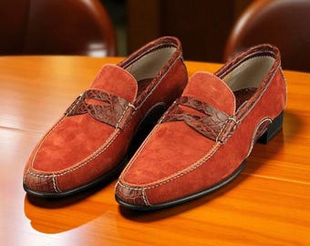 Könnte beinhalten: Ein Paar rostorangefarbene Wildleder-Loafer mit braunen Kroko-Leder-Akzenten. Die Schuhe haben weiße Nähte und einen niedrigen Absatz. Die Loafer sind auf einer Holzoberfläche ausgestellt.