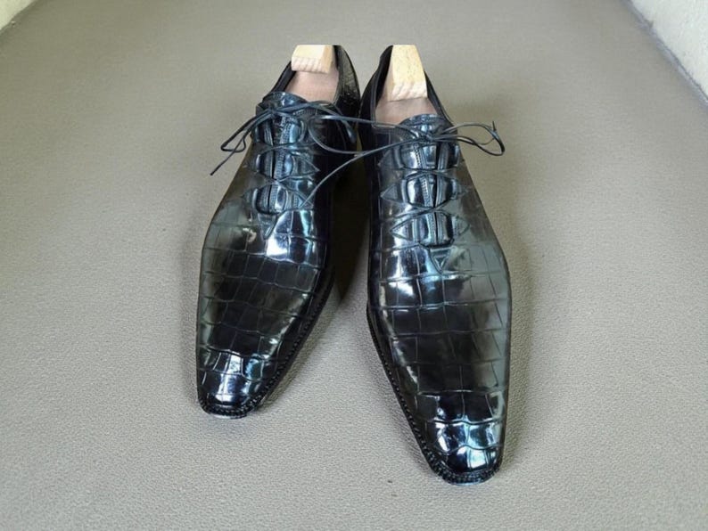 May include: A pair of black, glossy leather dress shoes with a textured, crocodile-like pattern. The shoes have laces and wooden shoe trees inside. The shoes are displayed on a neutral surface.