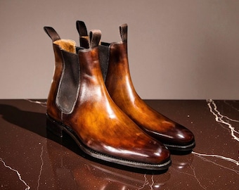 May include: A pair of brown leather Chelsea boots with a polished finish. The boots have elastic side panels, pull tabs, and a dark sole. The leather has a gradient effect, transitioning from light brown to a deep burgundy color.