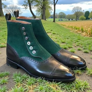 Puede incluir: Botines de dos tonos con la parte superior de ante verde y la puntera y el talón de cuero negro. Las botas tienen cierre de botón y están sobre un fondo de hierba.