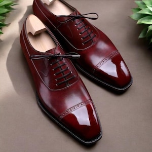 Pu&ograve; includere: Un paio di scarpe eleganti in pelle bordeaux con lacci neri. Le scarpe hanno una finitura lucida e cuciture decorative sulla punta. Le forme di legno sono all'interno delle scarpe. Le scarpe sono esposte su una superficie marrone.