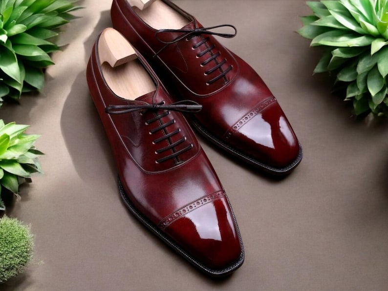 Pu&ograve; includere: Un paio di scarpe eleganti in pelle bordeaux con lacci neri. Le scarpe hanno una finitura lucida e cuciture decorative sulla punta. Le forme di legno sono all'interno delle scarpe. Le scarpe sono esposte su una superficie marrone.