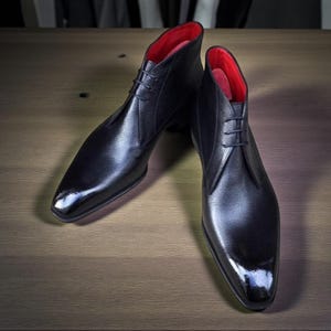 Puede incluir: Un par de botines de cuero negro con acabado brillante. Las botas tienen interior rojo y dise&ntilde;o con cordones. Los zapatos se exhiben sobre una superficie de madera.