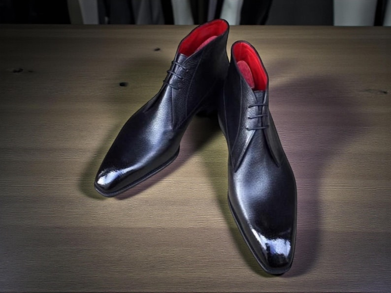 Puede incluir: Un par de botines de cuero negro con acabado brillante. Las botas tienen interior rojo y dise&ntilde;o con cordones. Los zapatos se exhiben sobre una superficie de madera.