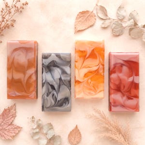May include: Four rectangular bars of soap arranged in a row. The first bar is a warm amber orange, the second is a cool grey with white swirls, the third is a bright golden yellow, and the fourth is a deep rose pink. Each bar has a smooth finish.