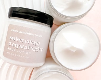 Può includere: Tre barattoli bianchi di lozione per il corpo con tappi neri. Il barattolo anteriore ha un'etichetta che recita "sweet clementine soaps sweet cream & crystal sugar BODY LOTION NET WT 5 OZ | 141.9"