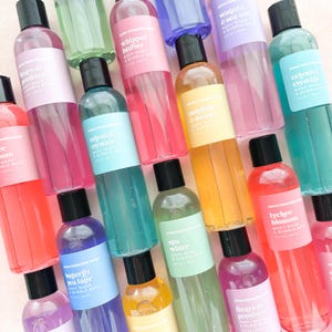 May include: A collection of colorful body wash and bubble bath bottles with black caps. The bottles are labeled with various scents like "Celestial Crystals," "Whipped Berries," and "Lychee Blossom." The background is a light pink.