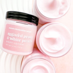 May include: Three jars of body lotion with pink lids and labels. The label on the front jar reads "sweet clementine soaps sugared peony & white peach BODY LOTION NET WT 5 OZ | 141g".