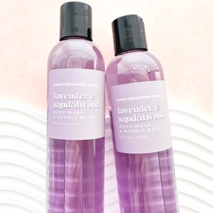 May include: Two clear plastic bottles filled with purple lavender and sandalwood body wash and bubble bath. Each bottle features a black cap and a light purple label with white text reading sweet clementine soaps lavender & sandalwood 9 fl oz | 266 ml.