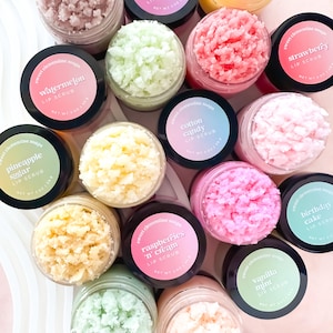 May include: An overhead shot of several small jars of lip scrub. The jars are filled with colorful, textured scrubs in various shades of pink, green, yellow, and white. Each jar has a black lid with a label indicating the flavor, such as "Watermelon" or "Strawberry".