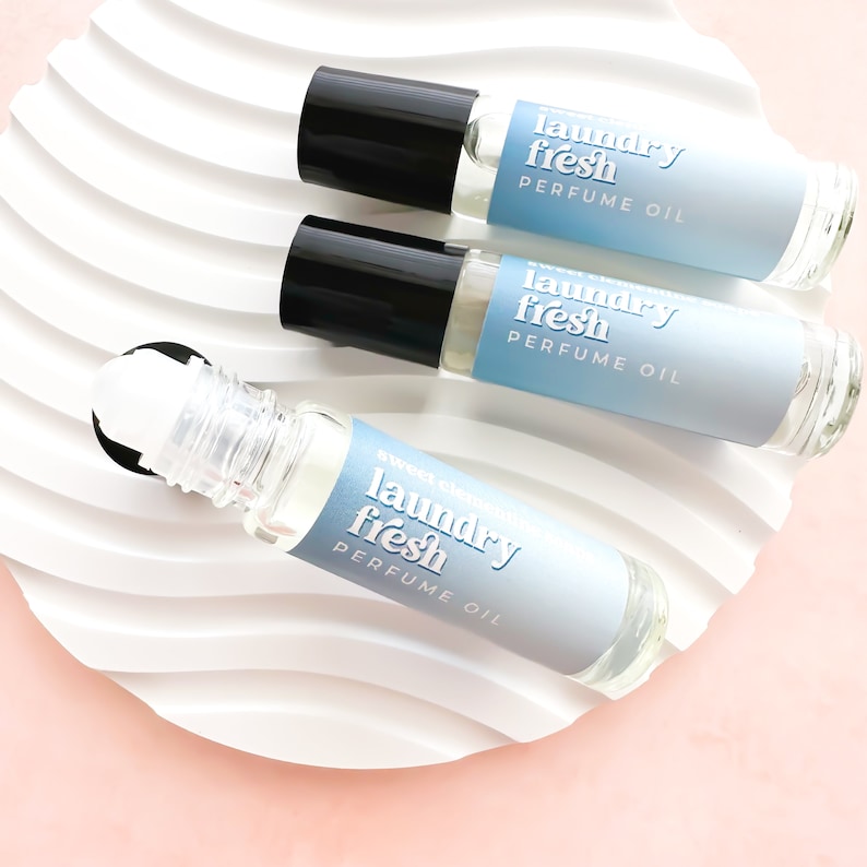 May include: Three glass roller bottles with black caps and labels that say "laundry fresh perfume oil". The bottles are on a white wavy surface.