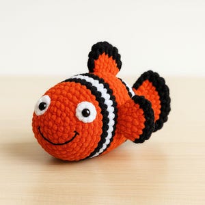 Könnte beinhalten: Ein gehäkelter orangefarbener Clownfisch mit schwarzen und weißen Streifen und Flossen. Der Fisch hat große weiße Augen mit schwarzen Pupillen und einen lächelnden Mund. Das handgefertigte Spielzeug liegt auf einer hellbraunen Holzoberfläche.