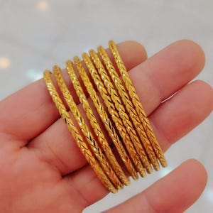 Puede incluir: Una colección de ocho pulseras doradas con un diseño trenzado texturizado. Las pulseras se sostienen en una mano, mostrando sus intrincados detalles y su acabado brillante. Las joyas se exhiben sobre un fondo suave y neutro.