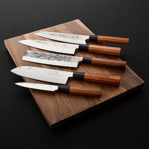 May include: Five stainless steel kitchen knives with wooden handles are arranged on a wooden cutting board. The blades have a textured finish. The handles are a light brown color and have a black bolster. The cutting board is a dark brown color.