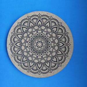 May include: Circular cork coaster with a black mandala design. The coaster has a symmetrical floral pattern with detailed lines and shapes. The coaster is a natural tan colour, set against a blue background.