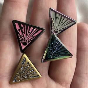 以下が含まれることがあります： サンバーストデザインの三角形のエナメルピン4個。ピンは黒、ピンク、虹色、金色の配色です。サンバーストのデザインは白、ピンク、シルバーです。ピンはおそらくジャケットやバックパック用です。