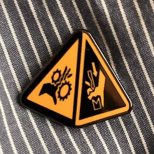 May include: A black and yellow triangular pin with safety symbols. The pin features a hazard symbol with a gear and a hand with a falling object. The pin is set against a blue and white striped fabric.