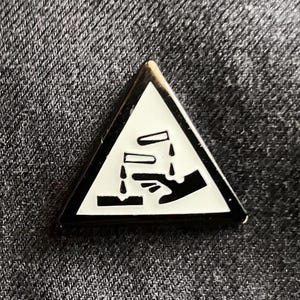 Könnte beinhalten: Ein kleiner, dreieckiger Ansteckpin mit einem weißen Gefahrensymbol für ätzende Stoffe. Der Pin hat einen goldfarbenen Rand und befindet sich auf einem dunkelgrauen Stoffhintergrund.