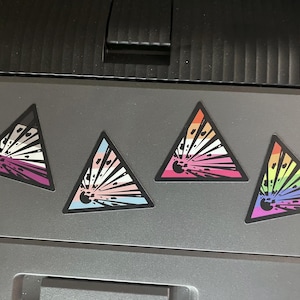 Può includere: Quattro adesivi triangolari con bordo nero. Ogni adesivo presenta un design a scoppio con una diversa combinazione di colori: viola, rosa, arancione e arcobaleno. Gli adesivi sono posizionati su una superficie grigia.