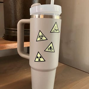 K&ouml;nnte beinhalten: Ein hellgrauer isolierter Becher mit wei&szlig;em Deckel und Strohhalm. Der Becher hat einen Griff und ist mit gelben Gefahrensymbol-Aufklebern verziert. Der Becher steht auf einem Holzregal.