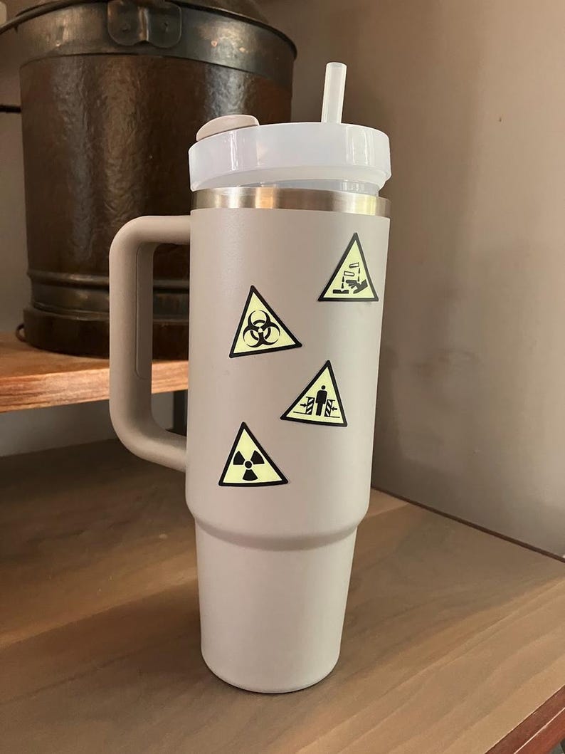 K&ouml;nnte beinhalten: Ein hellgrauer isolierter Becher mit wei&szlig;em Deckel und Strohhalm. Der Becher hat einen Griff und ist mit gelben Gefahrensymbol-Aufklebern verziert. Der Becher steht auf einem Holzregal.