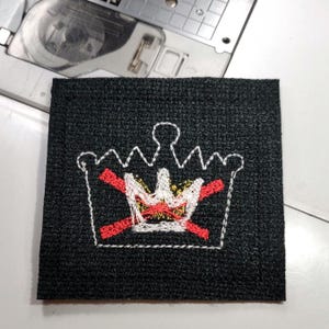 May include: Square black fabric patch with an embroidered crown design. The crown is white with red and gold accents, and two red lines cross through the design. The patch is likely for sewing or crafting.