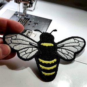 May include: Embroidered bee patch with black body, yellow stripes, and silver wings. The patch is held in front of a sewing machine, showcasing the detail of the insect design. This patch is ideal for adding to clothing or accessories.