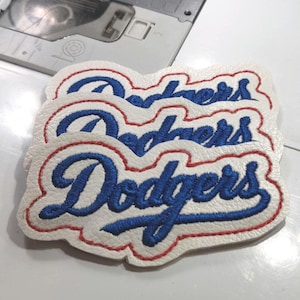 May include: Three white patches with the word "Dodgers" embroidered in blue script with a red border. The patches are stacked on top of each other. The background includes a sewing machine.