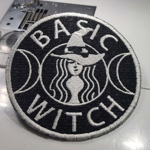May include: Round black and white embroidered patch with the words "BASIC WITCH" and a witch illustration. The witch has long flowing hair and a pointed hat. The patch is surrounded by a silver border and two crescent moons.