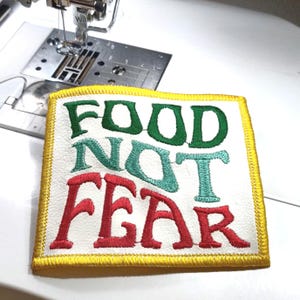 May include: Embroidered patch with the words "FOOD NOT FEAR" in green, teal, and red. The patch has a white background with a yellow border. The patch is on a white surface near a sewing machine.