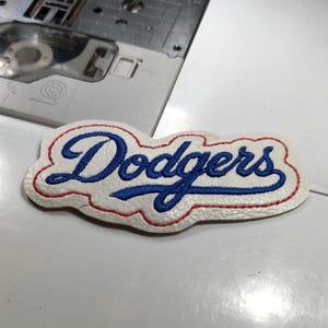 May include: A white patch with the word "Dodgers" embroidered in blue script, outlined in red. The patch has a textured appearance and is likely made of fabric. The patch is on a white surface.