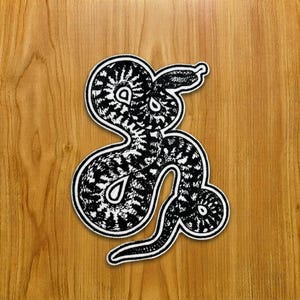 Op de afbeelding: Een zwart-witte geborduurde patch met een ontwerp van een kronkelende slang met ingewikkelde schubpatronen en een witte rand. De slang is afgebeeld in een gestileerde, grafische illustratiestijl met een zichtbare tong en een gebogen lichaamsvorm.