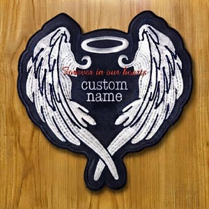 May include: A dark navy blue embroidered patch shaped like a heart with two large white angel wings and a halo at the top. The patch features the red text "forever in our hearts" and the white text "custom name" centred in the middle.