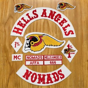 Puede incluir: Una colección de parches bordados con el texto HELLS ANGELS y NOMADS en rojo sobre fondo blanco. Incluye un gran emblema de calavera alada, dos parches en forma de diamante con el texto 1% y etiquetas rectangulares que dicen MC, AFFA y 666.