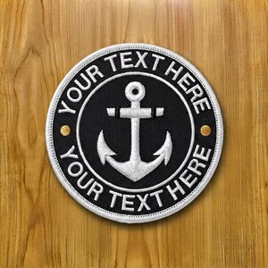 以下が含まれることがあります： 黒い背景の中央に白い錨が刺繍された円形のワッペン。白い縁取りには太字のサンセリフ体で「YOUR TEXT HERE」という文字が記されており、2つの小さな金色の円形スタッドがアクセントとして配置されています。