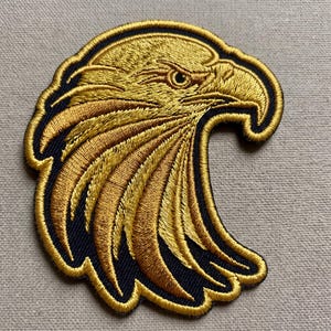 May include: An embroidered patch featuring the profile of a bald eagle head. The design is rendered in metallic gold thread against a dark navy blue background, with a thick gold border outlining the shape of the eagle's head and feathers.
