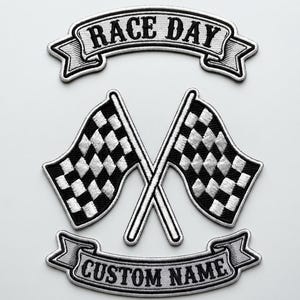 May include: A set of three embroidered patches featuring a black and white checkered flag design crossed in the center, with a top banner reading RACE DAY and a bottom banner reading CUSTOM NAME, all with a white border and black stitching.