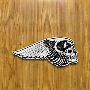 Könnte beinhalten: Ein schwarz-weiß bestickter Aufnäher mit einem stilisierten Totenkopf, der einen Helm mit einem großen, nach links ausladenden Flügel trägt. Das Design verwendet kontrastreiches schwarzes Garn auf weißem Grund für die Skelettdetails.