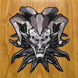May include: A black and white embroidered patch featuring a jester head with a pointed hat, sharp facial features, and a wide grin with visible teeth. The design is outlined in a thick black border, showcasing detailed shading and high contrast.