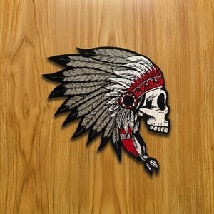 Puede incluir: Un parche bordado que muestra una calavera con un tocado de plumas de estilo tradicional. La calavera es de color blanco roto con sombreado negro, mientras que el tocado presenta plumas negras, grises y blancas con una banda roja y detalles rojos.