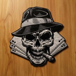 Könnte beinhalten: Ein schwarz-weiß bestickter Aufnäher, der einen Totenkopf mit Fedora-Hut zeigt, welcher eine Zigarre raucht. Der Totenkopf ist über einem Fächer aus Spielkarten positioniert, darunter der Pik-König, die Pik-Dame und eine Pik-Zehn.