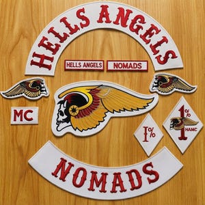 Puede incluir: Una colección de parches bordados que incluye un parche blanco grande en forma de arco con el texto rojo HELLS ANGELS, un arco inferior a juego con el texto rojo NOMADS, un emblema central de calavera alada, parches de calaveras aladas más pequeños y parches en forma de diamante 1%.