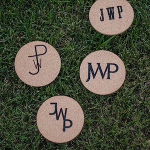 May include: Four round cork coasters with black monogram initials are arranged on green grass. The initials are JWP, P with a J, WMP, and JP with a W. The coasters are a natural cork color.