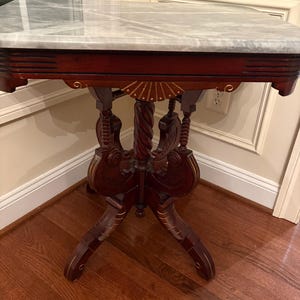 May include: A marble-topped side table with a dark wood frame. The table features ornate carvings and a pedestal base. The marble top has gray veining. The table is positioned in a corner of a room with hardwood flooring.