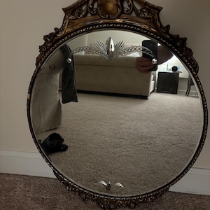 May include: An oval-shaped mirror with an ornate, gold-colored frame. The frame features intricate scrollwork and a decorative top. The mirror's surface reflects the room, and it has a decorative etched design at the top and bottom.