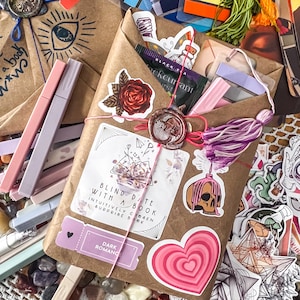 May include: A brown paper package wrapped with twine and decorated with a variety of stickers, including a red rose, a book, a heart, and a skull. The package is labeled "Blind Date with a Book" and "Dark Romance".