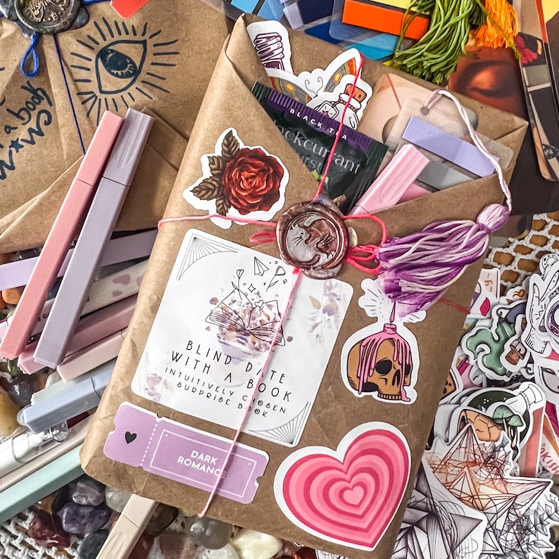 May include: A brown paper package wrapped with twine and decorated with a variety of stickers, including a red rose, a book, a heart, and a skull. The package is labeled "Blind Date with a Book" and "Dark Romance".